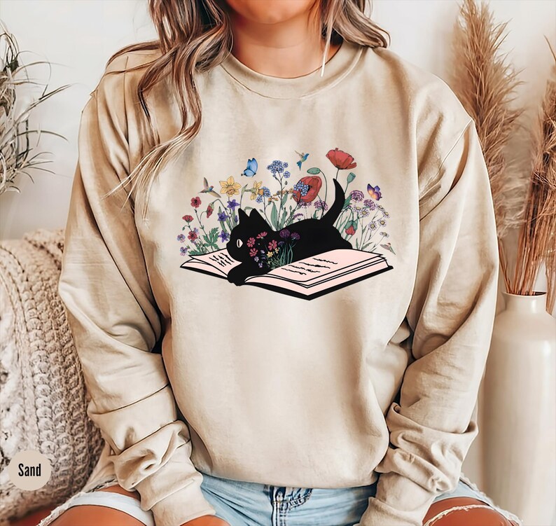 May include: A beige sweatshirt with a black cat reading a book surrounded by flowers. The cat is lying on the open book. The flowers are in shades of pink, purple, blue, and yellow.