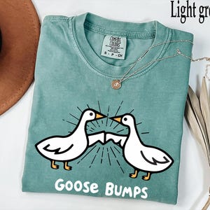 May include: A light green t-shirt featuring a white goose design and the text "Goose Bumps".