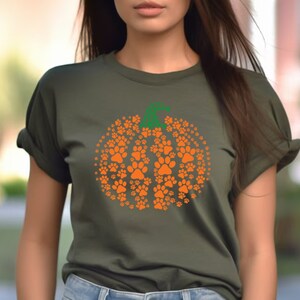 Peut inclure: T-shirt vert olive avec un motif citrouille orange compos&eacute; d'empreintes de pattes. La citrouille a une tige verte. Le t-shirt est &agrave; col rond et a des manches retrouss&eacute;es. Le motif est centr&eacute; sur le devant.