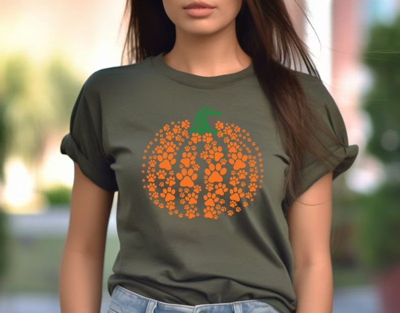 Peut inclure: T-shirt vert olive avec un motif citrouille orange compos&eacute; d'empreintes de pattes. La citrouille a une tige verte. Le t-shirt est &agrave; col rond et a des manches retrouss&eacute;es. Le motif est centr&eacute; sur le devant.