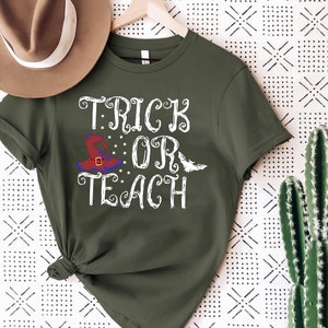 Puede incluir: Camiseta verde oliva con las palabras "TRICK OR TEACH" en escritura blanca. Se presenta un sombrero de bruja con detalles rojos, azules y morados, junto con un murci&eacute;lago blanco. La camiseta est&aacute; colgada en una percha de madera.