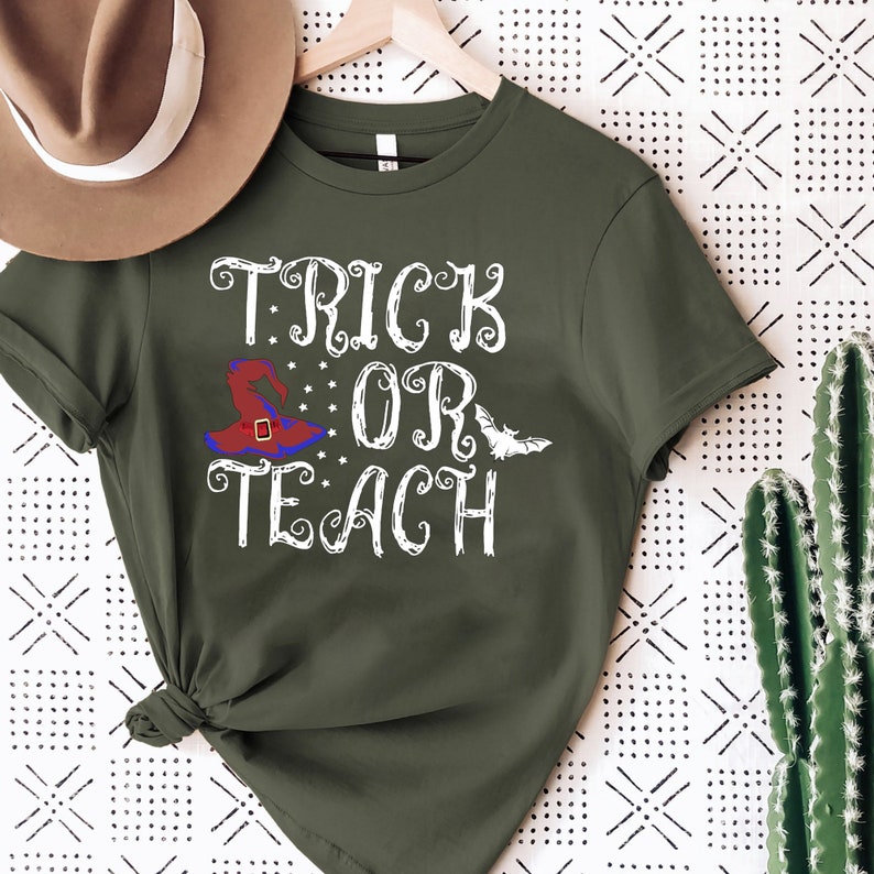 Puede incluir: Camiseta verde oliva con las palabras "TRICK OR TEACH" en escritura blanca. Se presenta un sombrero de bruja con detalles rojos, azules y morados, junto con un murci&eacute;lago blanco. La camiseta est&aacute; colgada en una percha de madera.