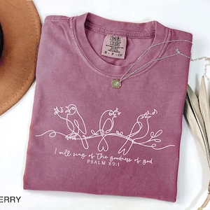May include: A berry-coloured t-shirt with a crew neck and short sleeves. The shirt features a white line drawing of three birds on a branch, with the text "I will sing of the goodness of god, PSALM 89:1." A silver necklace with a pendant is draped on the shirt.