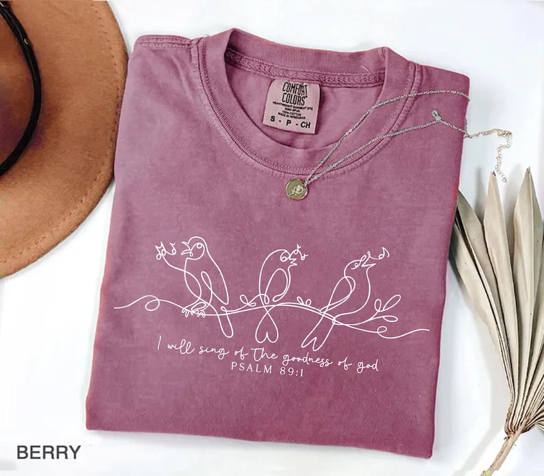 May include: A berry-coloured t-shirt with a crew neck and short sleeves. The shirt features a white line drawing of three birds on a branch, with the text "I will sing of the goodness of god, PSALM 89:1." A silver necklace with a pendant is draped on the shirt.