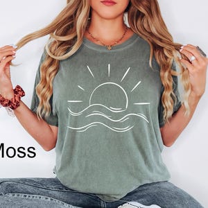 Puede incluir: Una camiseta verde musgo con un gr&aacute;fico blanco de un sol poni&eacute;ndose sobre las olas del oc&eacute;ano. La palabra "Moss" est&aacute; impresa en negro en la parte inferior izquierda. La camiseta es de material suave y tiene un corte holgado.