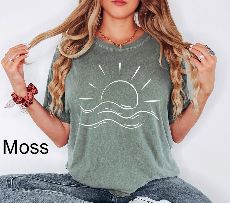 Puede incluir: Una camiseta verde musgo con un gr&aacute;fico blanco de un sol poni&eacute;ndose sobre las olas del oc&eacute;ano. La palabra "Moss" est&aacute; impresa en negro en la parte inferior izquierda. La camiseta es de material suave y tiene un corte holgado.