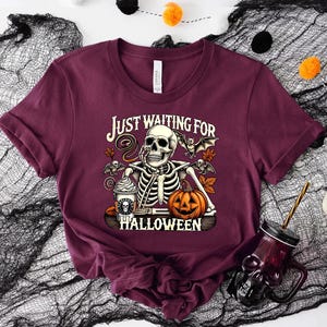 May include: A maroon t-shirt with a skeleton holding a pumpkin and a coffee cup. The text on the shirt reads "Just Waiting for Halloween".
