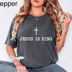 K&ouml;nnte beinhalten: Dunkelgraues T-Shirt mit dem wei&szlig;en Text "JESUS IS KING" und einem wei&szlig;en Kreuz dar&uuml;ber. Das Shirt hat einen Rundhalsausschnitt und kurze &Auml;rmel. Das Wort "Pepper" befindet sich in der oberen linken Ecke.