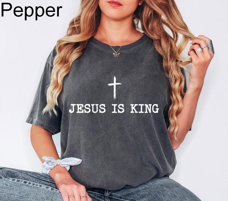 K&ouml;nnte beinhalten: Dunkelgraues T-Shirt mit dem wei&szlig;en Text "JESUS IS KING" und einem wei&szlig;en Kreuz dar&uuml;ber. Das Shirt hat einen Rundhalsausschnitt und kurze &Auml;rmel. Das Wort "Pepper" befindet sich in der oberen linken Ecke.