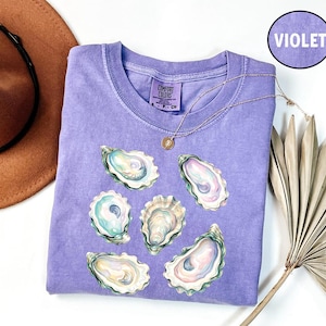 May include: A violet short-sleeve t-shirt featuring a graphic print of five oyster shells in shades of blue, green, and pink. The shirt has a Comfort Colors brand label at the inner neckline and a circular violet icon labeled VIOLET.