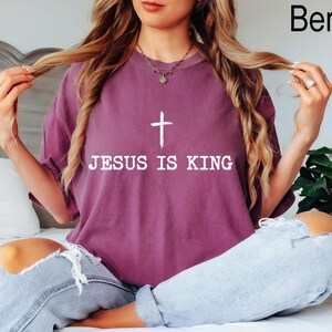 K&ouml;nnte beinhalten: Beerenfarbenes T-Shirt mit dem Text "JESUS IS KING" und einer wei&szlig;en Kreuzgrafik. Das Shirt hat eine lockere Passform und einen Rundhalsausschnitt. Das Wort "Berry" ist oben rechts aufgedruckt.