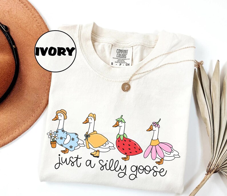 May include: Ivory-colored sweatshirt featuring a whimsical illustration of four geese dressed in various outfits, including a floral dress, a yellow cape, and a strawberry costume. The text "just a silly goose" is written below the geese.