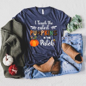 May include: Navy blue t-shirt with the text "I Teach the Cutest Pumpkins in the Patch" and a pumpkin graphic. The shirt is paired with blue jeans, brown boots, and a green sweater. Decorative pumpkins are also present.