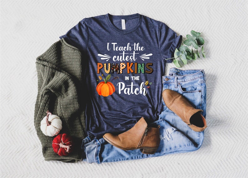 May include: Navy blue t-shirt with the text "I Teach the Cutest Pumpkins in the Patch" and a pumpkin graphic. The shirt is paired with blue jeans, brown boots, and a green sweater. Decorative pumpkins are also present.
