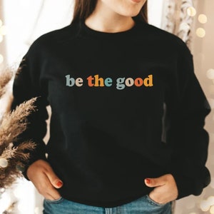 May include: A black sweatshirt with the text "be the good" in colorful letters. The letters are in a retro font style.