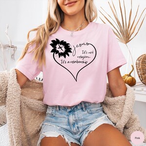 May include: A light pink t-shirt featuring a black graphic of a sunflower forming the top of a heart shape. The text inside the heart reads, "Jesus It's not religion... It's a relationship." The shirt has short sleeves and a crew neckline.