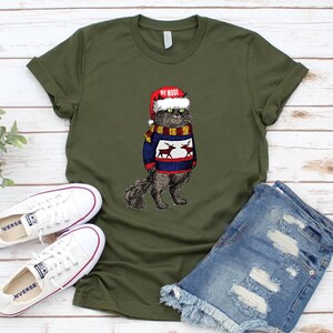 K&ouml;nnte beinhalten: Ein gr&uuml;nes T-Shirt mit einer schwarzen Katze, die einen roten Weihnachtsmann-Hut und einen blau-wei&szlig;en Pullover mit Rentieren tr&auml;gt. Der Text "MY MODE" steht auf dem Hut.