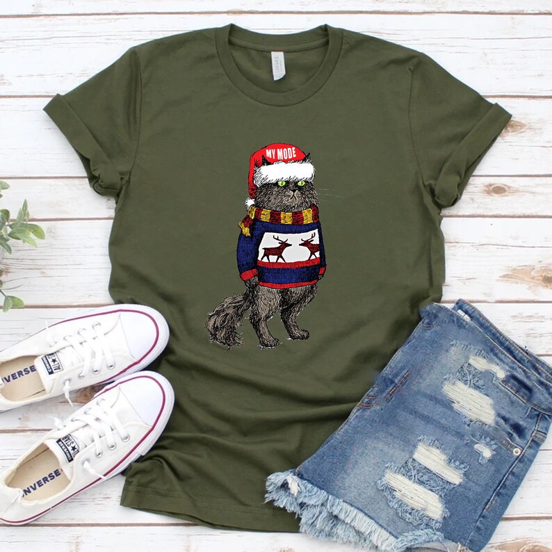 K&ouml;nnte beinhalten: Ein gr&uuml;nes T-Shirt mit einer schwarzen Katze, die einen roten Weihnachtsmann-Hut und einen blau-wei&szlig;en Pullover mit Rentieren tr&auml;gt. Der Text "MY MODE" steht auf dem Hut.
