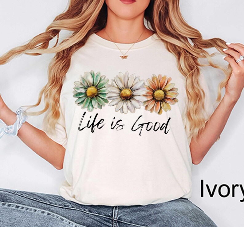 May include: Ivory-colored t-shirt featuring three daisy flowers in green, white, and orange with yellow centers. The phrase "Life is Good" is printed in black script below the flowers. The shirt is a casual, everyday wear item.