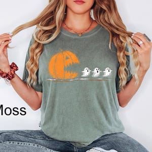 May include: Moss green t-shirt featuring a Halloween-themed graphic. The design includes an orange jack-o'-lantern with a wide open mouth, and three small white ghosts. The word "Moss" is printed in black on the bottom left of the shirt.