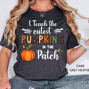May include: Dark grey heather t-shirt with the text "I Teach the cutest PUMPKINS in the Patch." The word "PUMPKINS" is in orange, green, and black. An orange pumpkin graphic is below the text. The shirt is short-sleeved.