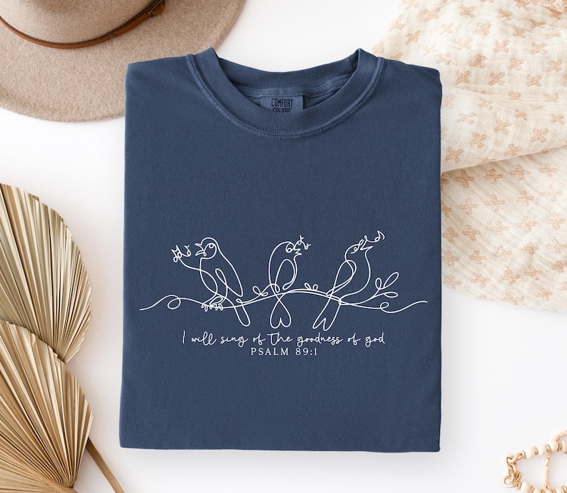 May include: Navy blue sweatshirt featuring a white line drawing of three birds on a branch. The text reads "I will sing of the goodness of God, Psalm 89:1". The sweatshirt is folded neatly.