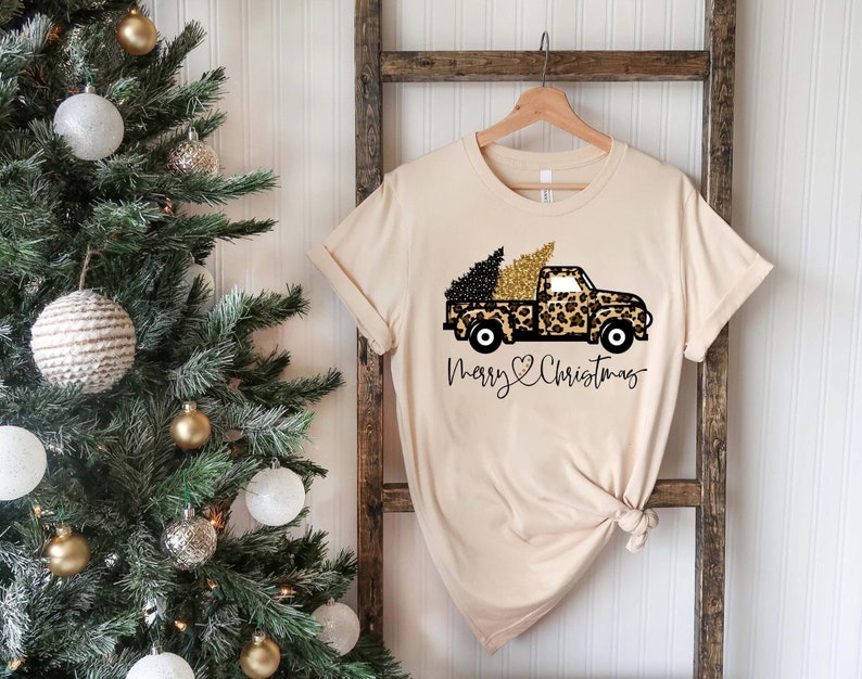 Op de afbeelding: Een beige T-shirt met een zwarte en gouden luipaardprint truck die een kerstboom vervoert. De tekst "Merry Christmas" is onder de truck gedrukt met een hartje.