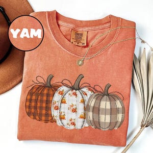 Può includere: T-shirt arancione Comfort Colors con tre zucche illustrate con diversi motivi: a quadri, floreale e cucito. Una collana d'oro con un ciondolo è drappeggiata sulla maglietta. La parola "YAM" è visibile su un'etichetta circolare.