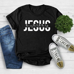 Pu&ograve; includere: Maglietta nera con testo bianco che recita "JESUS the way the truth the life". La maglietta &egrave; appoggiata su una superficie di legno bianco con un paio di scarpe Converse grigie e un paio di jeans blu.