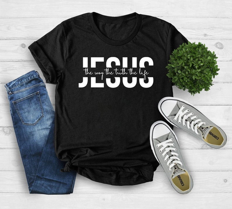 Pu&ograve; includere: Maglietta nera con testo bianco che recita "JESUS the way the truth the life". La maglietta &egrave; appoggiata su una superficie di legno bianco con un paio di scarpe Converse grigie e un paio di jeans blu.