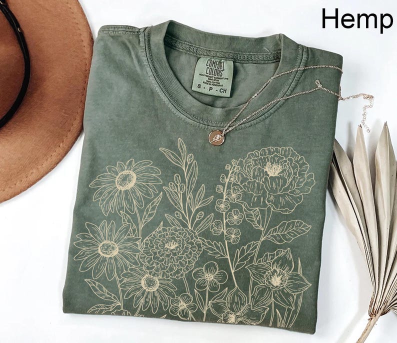 K&ouml;nnte beinhalten: Hanffarbenes T-Shirt mit floralem Design. Das Shirt hat einen Rundhalsausschnitt und kurze &Auml;rmel. Eine silberne Halskette mit Anh&auml;nger liegt auf dem Shirt. Das Wort "Hemp" ist oben rechts aufgedruckt.
