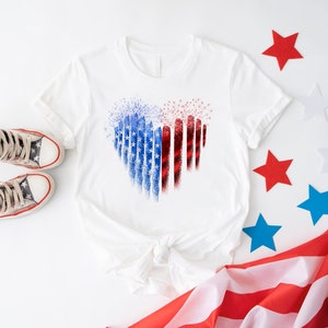 Pu&ograve; includere: T-shirt bianco con un design a forma di cuore nei colori rosso, bianco e blu, che ricorda la bandiera americana. L'immagine include scarpe da ginnastica con motivo a stelle e stelle decorative, evocando un tema patriottico.
