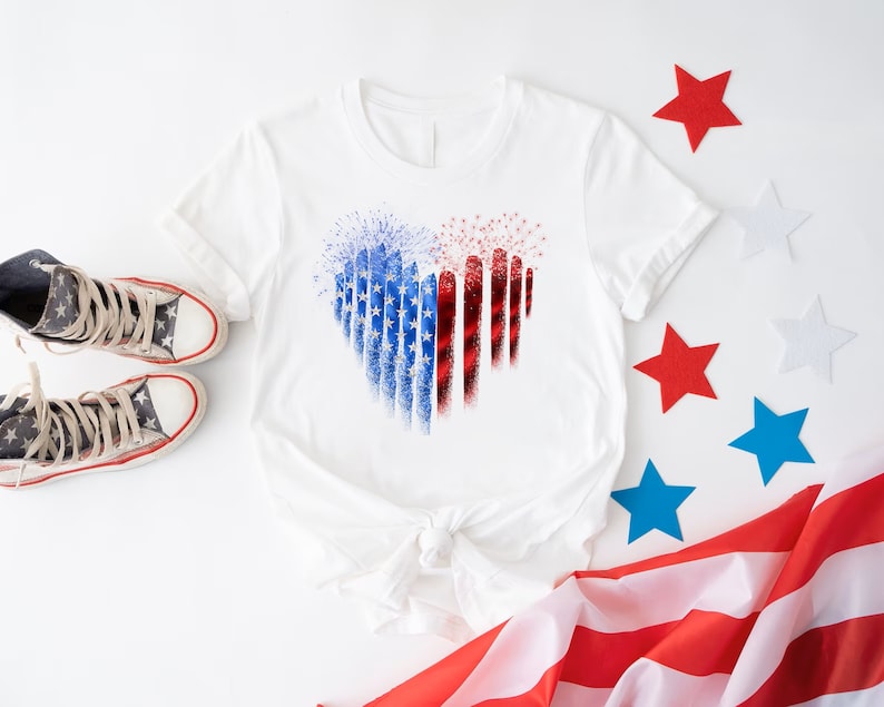 Pu&ograve; includere: T-shirt bianco con un design a forma di cuore nei colori rosso, bianco e blu, che ricorda la bandiera americana. L'immagine include scarpe da ginnastica con motivo a stelle e stelle decorative, evocando un tema patriottico.
