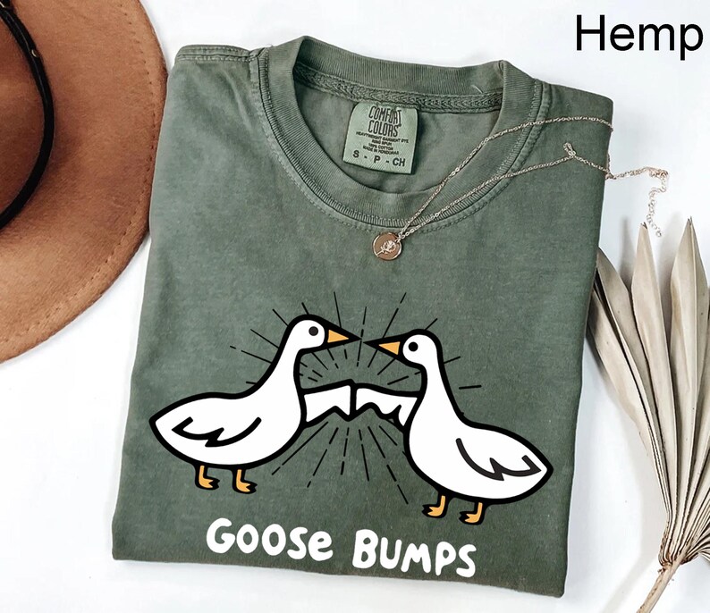 May include: A green t-shirt featuring a graphic of two geese facing each other with the text "Goose Bumps" printed below. The shirt is made from hemp.