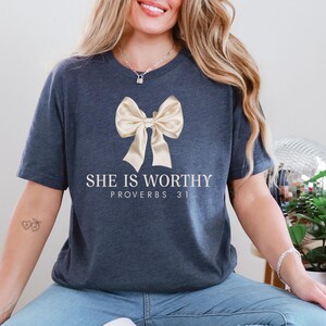 Puede incluir: Una camiseta azul marino con un lazo dorado y el texto "She is worthy Proverbs 31" impreso en la parte delantera.