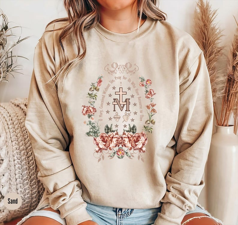 Puede incluir: Una sudadera beige con un dise&ntilde;o floral y el texto "Oh Mar&iacute;a, concebida sin pecado, ruega por nosotros que recurrimos a ti." El dise&ntilde;o incluye una cruz y las letras "M" y "A" entrelazadas.