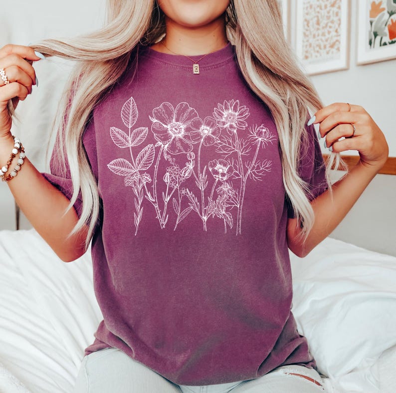 May include: A short-sleeved, plum-colored t-shirt featuring a white floral line drawing across the front. The design includes various flowers and foliage. The shirt is made of a soft material.