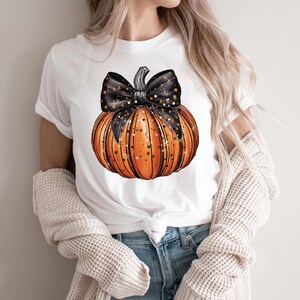 May include: White t-shirt featuring a graphic of an orange pumpkin with a black and white star patterned bow.