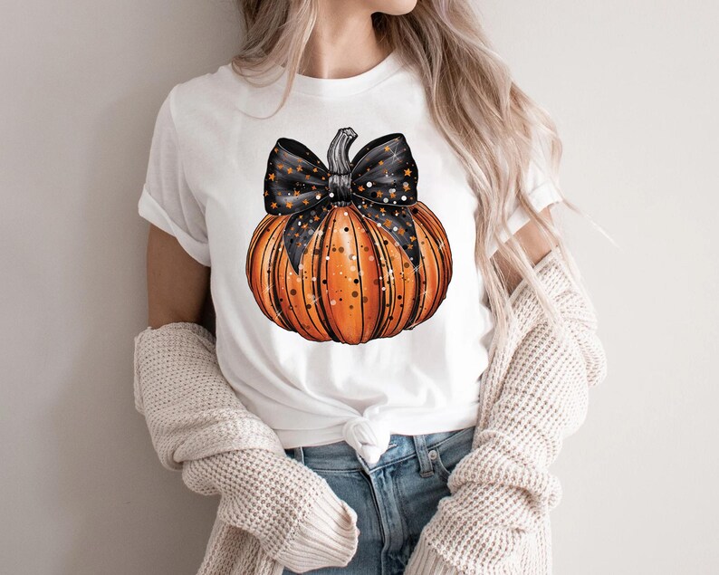 May include: White t-shirt featuring a graphic of an orange pumpkin with a black and white star patterned bow.