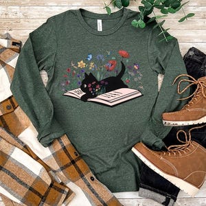 May include: A long-sleeved, heather forest green t-shirt with a black cat reading a book surrounded by flowers. The cat is sitting on the open book.