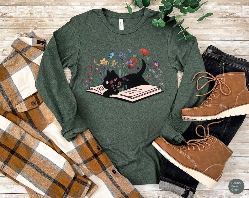 May include: A long-sleeved, heather forest green t-shirt with a black cat reading a book surrounded by flowers. The cat is sitting on the open book.