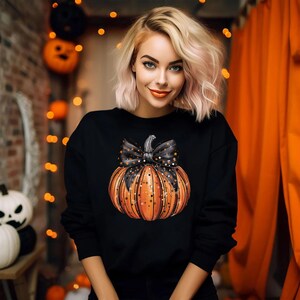 May include: A black sweatshirt featuring a large orange pumpkin with a black bow and glitter accents. The pumpkin is positioned centrally on the front of the sweatshirt.