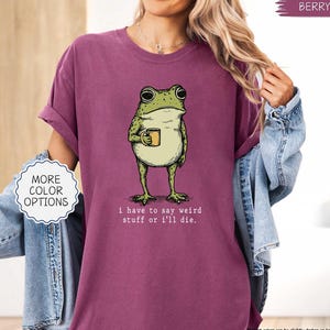 May include: Berry-colored t-shirt featuring a whimsical illustration of a frog holding a mug. The frog is green and beige, with the text "i have to say weird stuff or i'll die." A white circle with "MORE COLOR OPTIONS" is on the left.