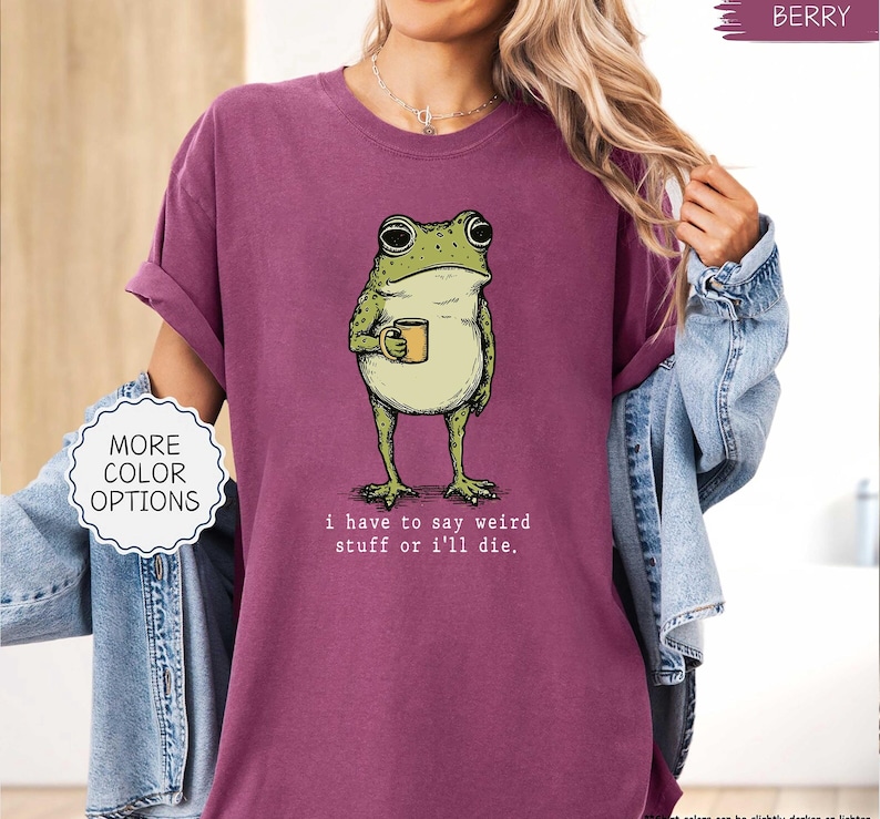 May include: Berry-colored t-shirt featuring a whimsical illustration of a frog holding a mug. The frog is green and beige, with the text "i have to say weird stuff or i'll die." A white circle with "MORE COLOR OPTIONS" is on the left.