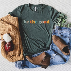 May include: A green t-shirt with the text "be the good" in colorful letters. The shirt is laid out on a white fuzzy surface with a brown sweater, blue jeans, and brown boots.