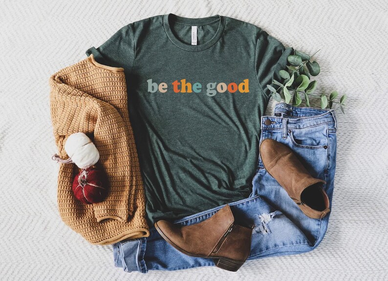 May include: A green t-shirt with the text "be the good" in colorful letters. The shirt is laid out on a white fuzzy surface with a brown sweater, blue jeans, and brown boots.