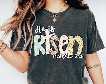 May include: Dark gray t-shirt with the words "He is Risen" in a colorful, patterned design. The word "Risen" features a cross design. The text "Matthew 28:6" is below the word "Risen".