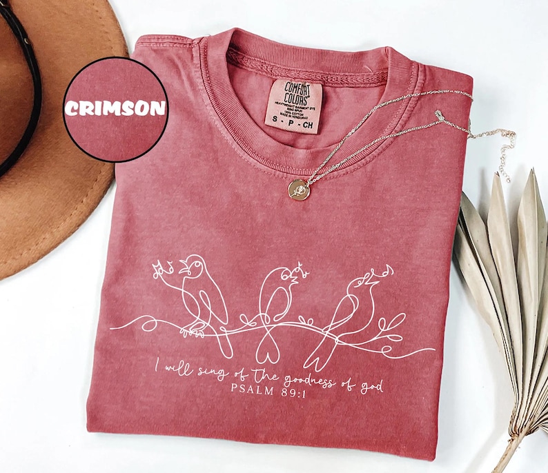 May include: Crimson t-shirt with a crew neck and short sleeves. The shirt features a white line drawing of three birds perched on a branch, with the text "I will sing of the goodness of god, Psalm 89:1". A silver necklace with a pendant is on the shirt.