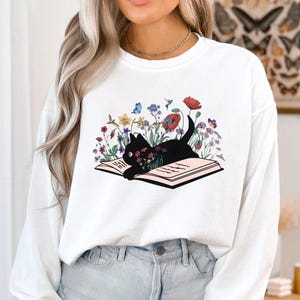 May include: White long-sleeve sweatshirt with a black cat reading a book surrounded by colorful flowers and butterflies.
