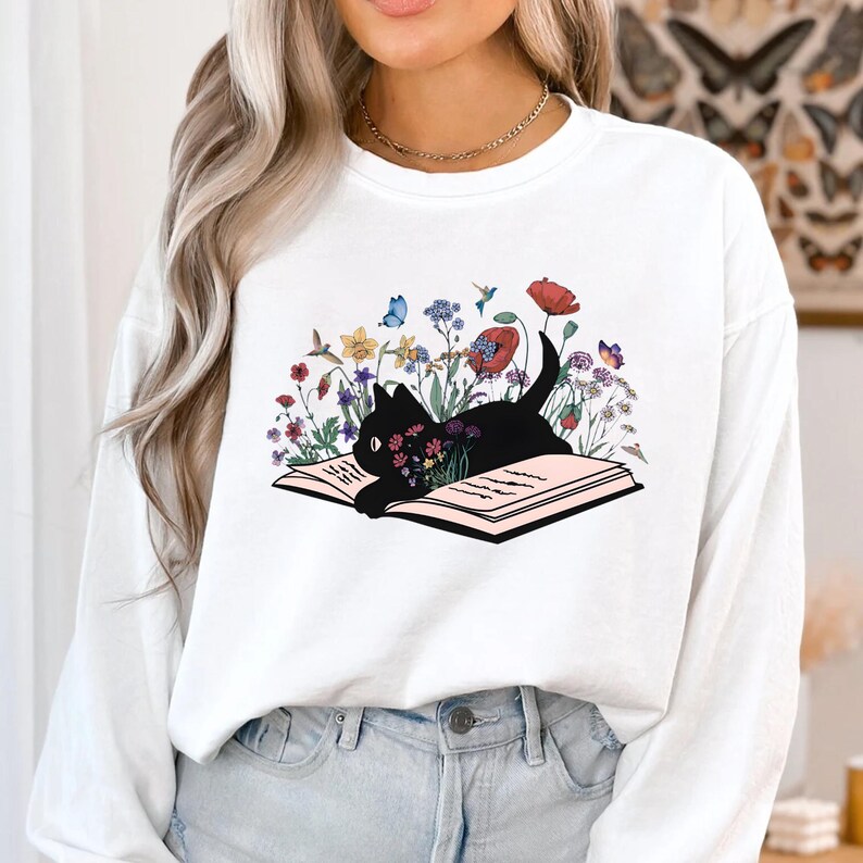 May include: White long-sleeve sweatshirt with a black cat reading a book surrounded by colorful flowers and butterflies.